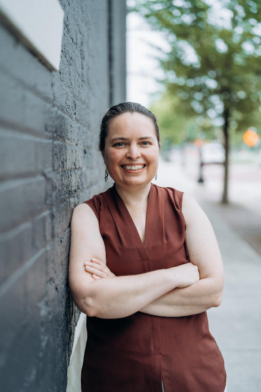I'm wearing a long sleeveless reddish-brown shirt and standing with folded arms by a blue-gray painted brick wall.